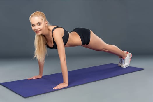 Young beautiful woman in fitness wear trained female body, doing push-ups on the mat. Look at the camera