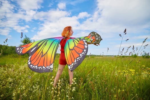 Adult girl with red hair and butterfly wings having fun and joy in meadow or field with grass, flowers on sunny summer day