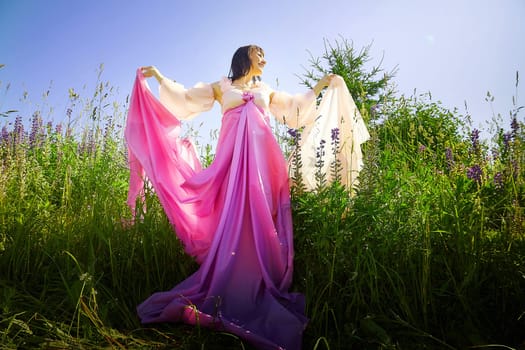 Beautiful girl in lush pink ball gown in green field during blooming of flowers and blue sky on background. Model posing on nature landscape as princess from fary tale