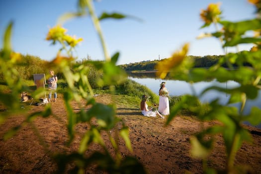 Adult female artist painting picture near water of river or lake in nature and girls in white sundress and flower wreath. Artist and models posing in holiday of Ivan Kupala in nature at sunset