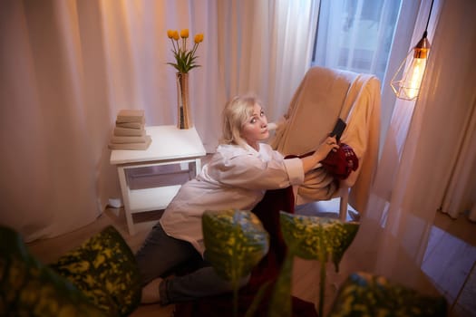 Adult mature woman of 40 or 60 years old with cell phone smartphone in casual dress white shirt and blue jeans in calm cozy evening atmosphere room. Interior with curtains and soft warm lamp