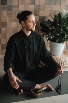Portrait of a young man in a black kimano sitting in a lotus position on a gym mat in the interior.