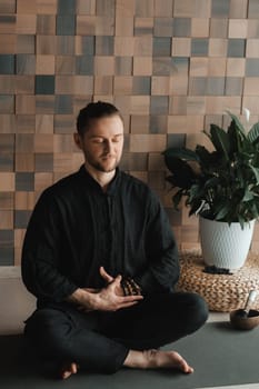 Portrait of a young man in a black kimano sitting in a lotus position on a gym mat in the interior.