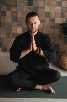 Portrait of a young man in a black kimano sitting in a lotus position on a gym mat in the interior.