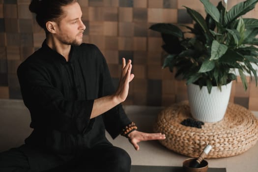 Portrait of a young man in a black kimano sitting in a lotus position on a gym mat in the interior.