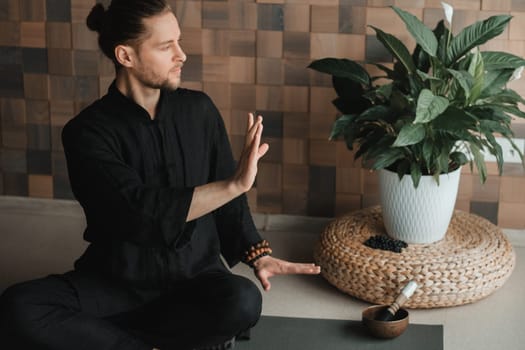 Portrait of a young man in a black kimano sitting in a lotus position on a gym mat in the interior.