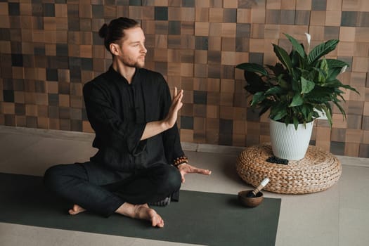 Portrait of a young man in a black kimano sitting in a lotus position on a gym mat in the interior.