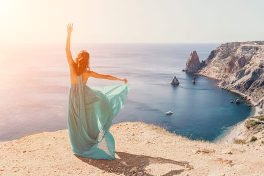 Side view a Young beautiful sensual woman in a red long dress posing on a rock high above the sea during sunrise. Girl on the nature on blue sky background. Fashion photo.