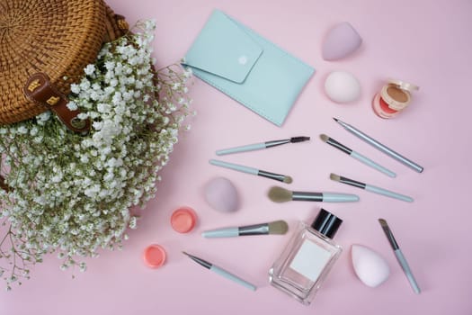 Makeup items are laid out on a pink background. There is a wicker bag with beautiful white gypsophila flowers on the table. Makeup items are laid out on the table.