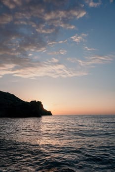 A red burning sunset with the silhouette of a cliff over the sea. Abstract nature summer or spring ocean sea background. Small waves on golden warm water surface with bokeh lights from sun.