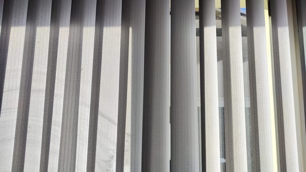Vertical fabric white blinds on the window with sun. Background and texture. Abstract pattern and frame. Stripes and lines of white-gray color