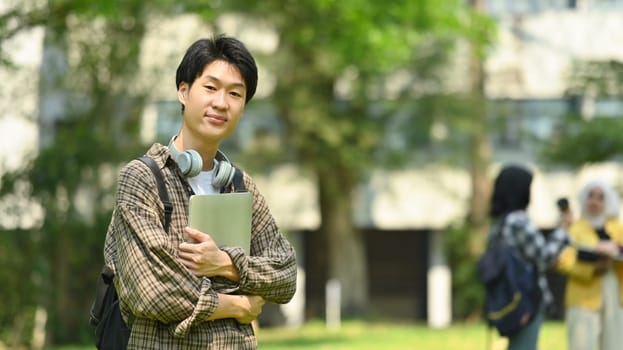 Asian man holding laptop standing at university campus with other students standing in background.