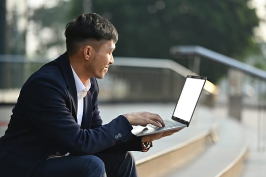 Image of young asian male worker using laptop on stairs in front of business center. Modern lifestyle, business, technology concept.