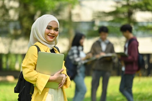 Smiling Asian muslim woman holding laptop standing at university campus with other students standing in background.