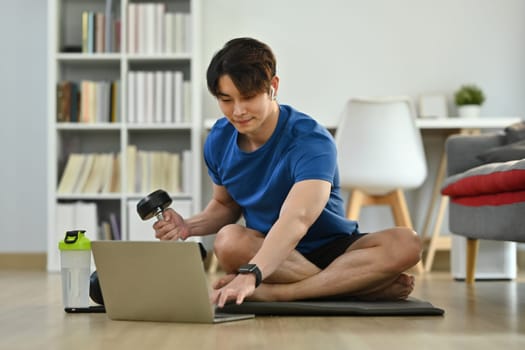 Full length of young man exercising with dumbbell and watching online tutorial on laptop. Healthy lifestyle and fitness concept.
