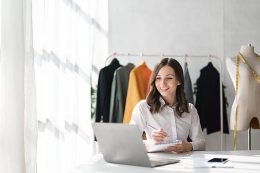 Young attractive female fashion designer sketching idea on paper at desk, working with a laptop at home.