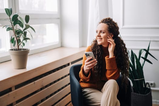 Young woman enjoying smartphone apps and texting while sitting on a chair. Mobile phone user: woman checking apps, messaging, and shopping from home. Relaxed woman browsing modern apps.