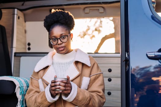 young african woman relaxing and enjoying a cup of a hot drink at sunset in a camper van, concept of van life and weekend getaway, copy space for text