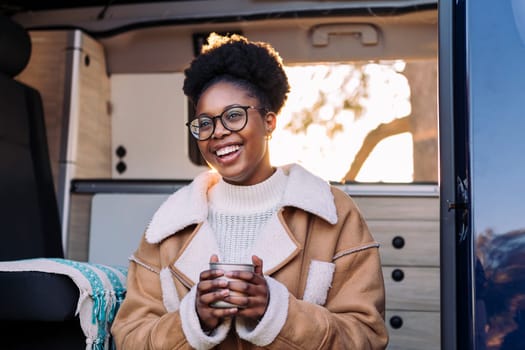 young african woman smiling happy at sunset relaxing in a camper van enjoying a cup of a hot drink, concept of van life and weekend getaway