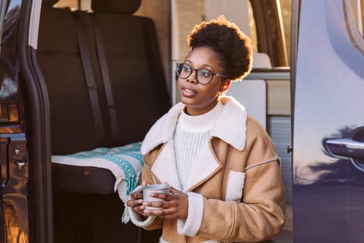 young african woman chilling and enjoying a mug of a hot drink at sunset in a camper van, concept of van life and weekend getaway, copy space for text