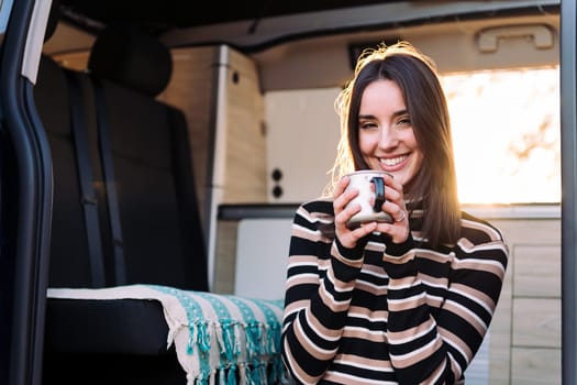 portrait of a young caucasian woman smiling happy in a camper van looking at camera with a mug in her hand, concept of van life and weekend getaway, copy space for text