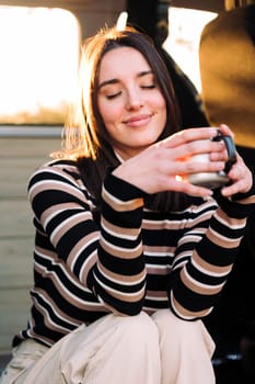 young caucasian woman chilling and enjoying a mug of a hot drink at sunset in a camper van, concept of van life and weekend getaway