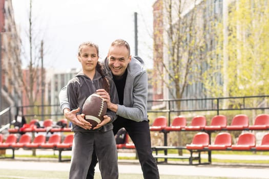 Handsome dad with his little cute daughter are having fun and playing American football on green grassy lawn.