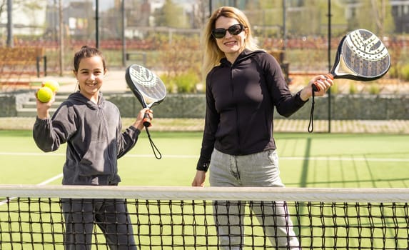 Full body of positive mother and daughter standing on court with padel rackets during break and looking at camera
