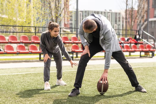 Handsome dad with his little cute daughter are having fun and playing American football on green grassy lawn.