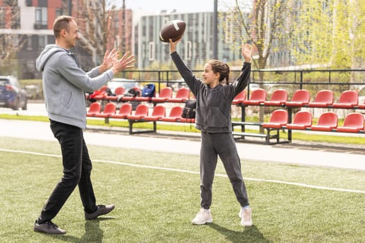 Handsome dad with his little cute daughter are having fun and playing American football on green grassy lawn.