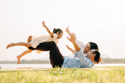 A joyful family moment in the park. Father holds his daughter up high as she smiles and flies like an airplane, while mother looks on with delight. Family fun at its best
