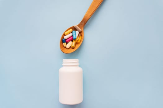 Various bright pills on a blue background lie in a wooden spoon along with a plastic bottle on the background. The concept of healthcare and evidence-based medicine, close-up