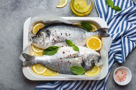 Raw fish dorado in casserole dish with ingredients lemon, fresh basil, bottle of olive oil on wooden cutting board with knife on rustic stone background top view, cooking healthy fish dorado concept