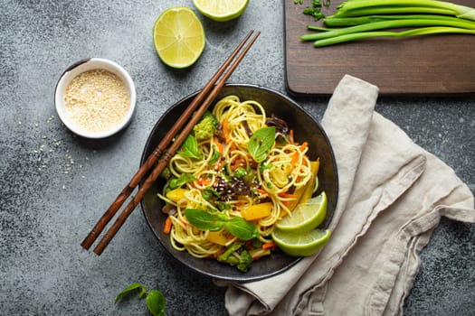 Asian vegetarian noodles with vegetables and lime in black rustic ceramic bowl, wooden chopsticks, cutting board with chopped green onion top view on stone background. Cooking noodles concept