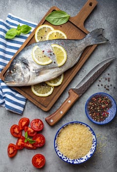 Raw fish dorado with ingredients lemon, fresh basil, cut cherry tomatoes, uncooked rice on wooden cutting board with knife on rustic stone background top view, cooking healthy fish dorado concept