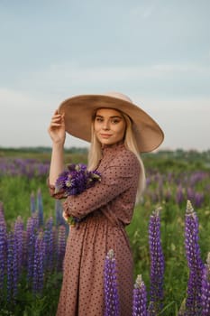 A beautiful woman in a straw hat walks in a field with purple flowers. A walk in nature in the lupin field.