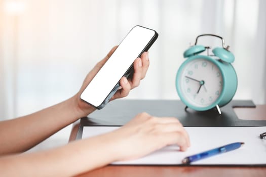 Woman's hand using a smartphone with blank white screen at work with desk accessories such as notepad, pen and alarm clock for the concept of business, communication and media technology.