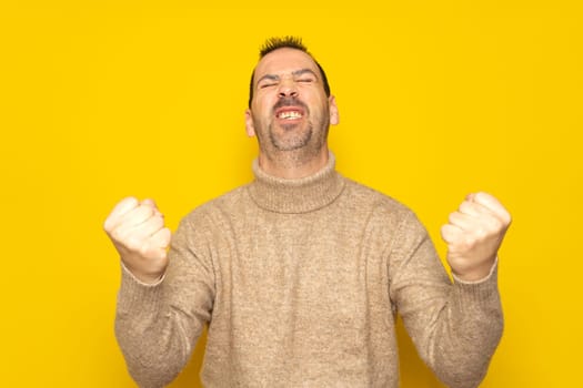 Bearded Hispanic man in his 40s wearing a beige turtleneck energetically celebrating a big win with raised fists, isolated on yellow studio background