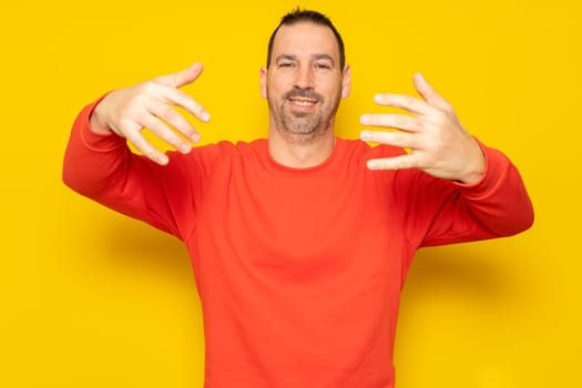 Smiling bearded hispanic man 40s wearing casual pullover posing stretching hands for hug while noticing someone looking at camera isolated on yellow background studio portrait