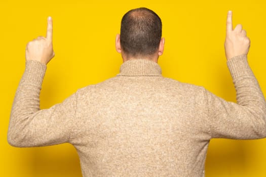 Hispanic man in casual clothing posing backwards pointing up with his index fingers, isolated on yellow studio background