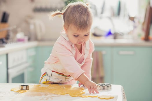 Adorable baby in an apron makes cookies from dough in the kitchen.