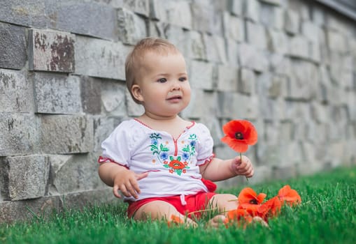 baby in embroidered shirt sitting on a lawn with flowers.