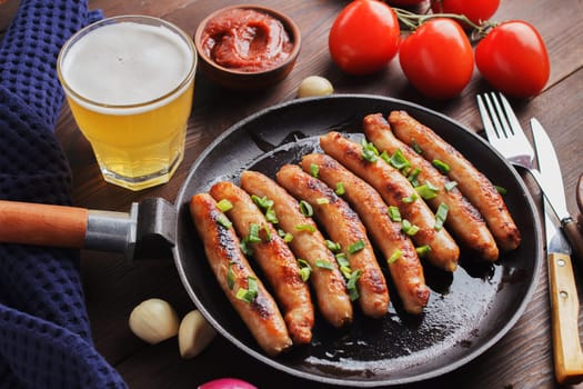 Delicious sausages cooked in a frying pan with vegetables, various sauces and a glass of beer on a wooden table.