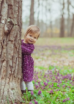 blond baby in a colored dress peeps out from behind a tree in the forest