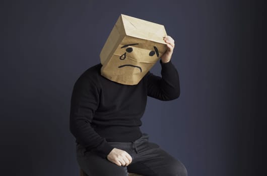 A sad man in a black turtleneck with a bag on his head, with a drawn crying emoticon, holds his head and cries. Emotions and gestures.