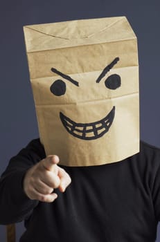 A man in a black turtleneck with a paper bag with an angry emoticon on his head shows his hand pointing forward. Emotions and anger. Vertical frame.