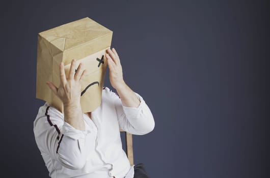 A sad man in a white shirt with a bag on his head, with a drawn sad emoticon is afraid, closes his eyes with his hands. Emotions and gestures.