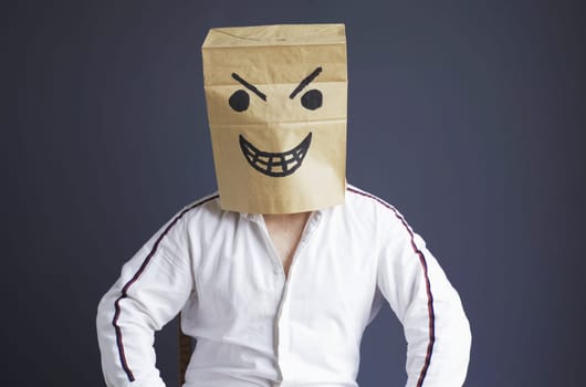 A man sits on a chair with a paper bag on his head, with a drawn angry smiley. Emotions and gestures.
