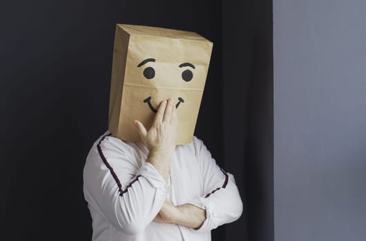A man in a white shirt with a paper bag on his head, with a painted smiley face, sends an air kiss.