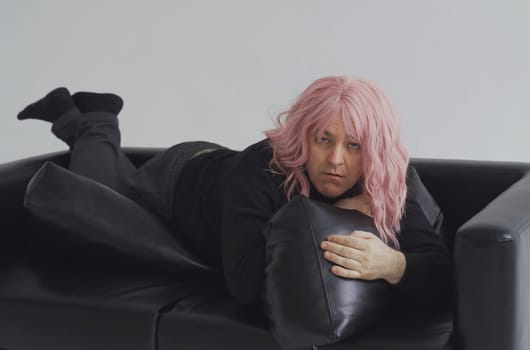 A drunk man in a pink wig lies on the couch, wakes up, raises his head and straightens his hair.